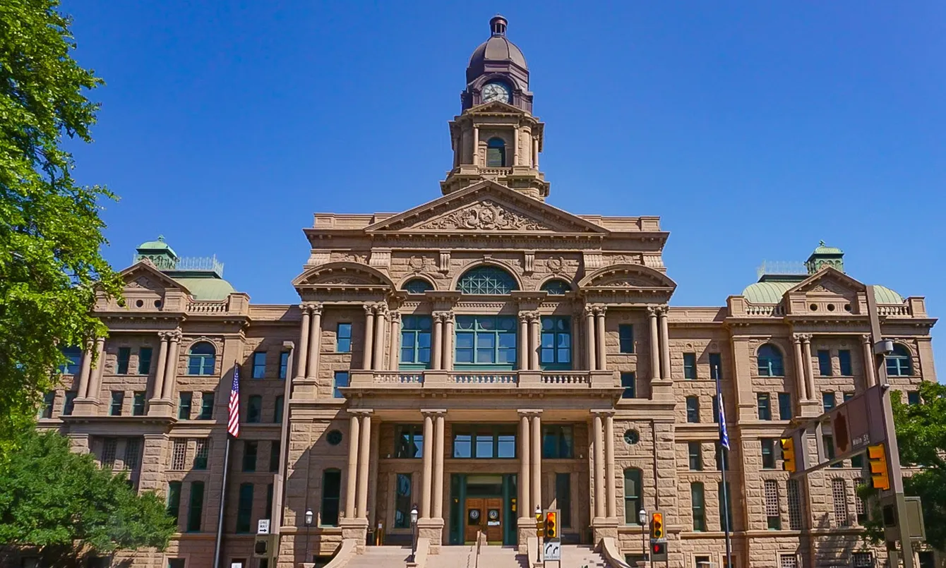 Fort Worth, TX Courthouse. Beige stone building with clock tower and US flag under a blue sky.